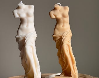 May include: Two sculpted candles in the shape of the Venus de Milo, one white and one beige, on a wooden surface with a white ribbon that says "Etoile" on it.