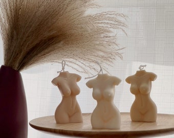 May include: Three beige torso-shaped candles with wicks sit on a wooden table. A burgundy vase with tall, dried pampas grass is in the background. The candles are in different shades of beige.