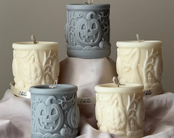 Puede incluir: Cinco velas cilíndricas con diseños tallados de Halloween. Dos son grises y tres son blanquecinas. Cada vela presenta una calabaza, árboles y otros elementos espeluznantes. Las velas tienen mechas y están dispuestas sobre una superficie drapeada.