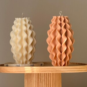 May include: Two decorative pillar candles with a wavy, textured design. One candle is a creamy off-white, the other is a warm peach color. Both candles have a silver wick and sit on a wooden tray.