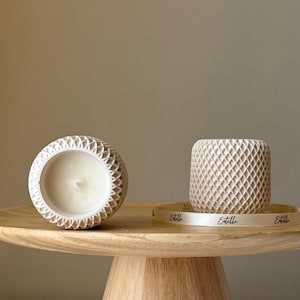 Op de afbeelding: Twee witte keramische kaarsenhouders met een geometrisch patroon. De kaarsenhouders staan op een houten voetstuk met een wit lint waarop "Etelle" staat.