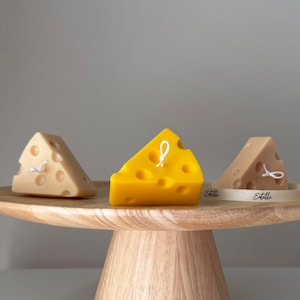May include: Three cheese-shaped candles on a wooden cake stand. The candles are yellow, beige, and brown. The candles are tied with white string and have a "Etelle" label.