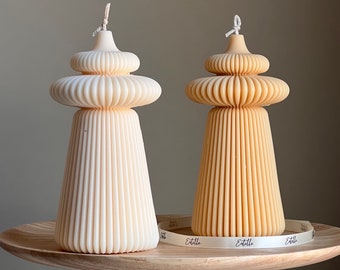 May include: Two decorative candles in the shape of a pagoda with a white and a beige color. The candles are on a wooden pedestal with a white ribbon that says "Etoile" on it.