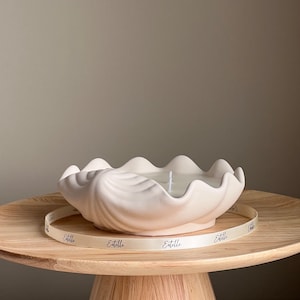 May include: White ceramic seashell candle holder with a white candle inside. The candle holder is sitting on a wooden table with a white ribbon that says "Etelle" on it.