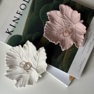 May include: Two leaf-shaped trinket dishes, one white and one pale pink, each holding rings. The dishes sit on a magazine with the word "KINFOLK" visible. The leaf dishes are a decorative and functional way to store jewelry.