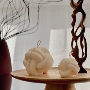 Peut inclure: Deux bougies blanches nouées sur une table en bois. La plus grande bougie a la forme d'un nœud et la plus petite bougie est une version plus petite du même nœud.
