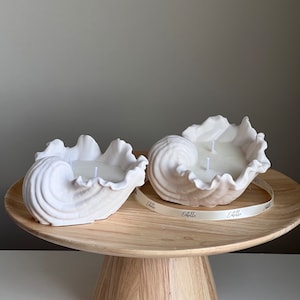 May include: Two white shell-shaped candles with two wicks each, sitting on a wooden pedestal with a white ribbon that says "Etelle" on it.