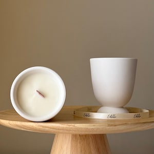 Op de afbeelding: Twee witte keramische kaarsenpotten met een houten lont. De kaarsenpotten staan op een houten voetstuk met een wit lint waarop "Etoile" staat.