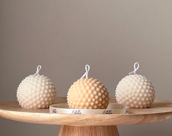 May include: Three round, textured candles in white and beige on a wooden pedestal. The candles are decorated with a white ribbon with the word "Estelle" printed on it.