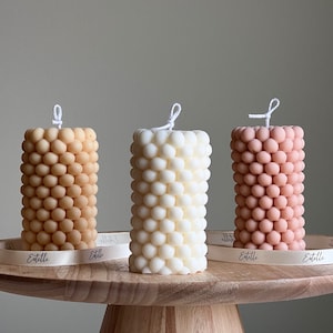 May include: Three pillar candles with a textured, bubble-like design. The candles are in shades of beige, white, and light pink. The candles are on a wooden pedestal.