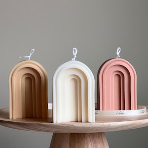 Könnte beinhalten: Drei bogenförmige Kerzen in verschiedenen Farben: Beige, Weiß und Rosa. Die Kerzen sind auf einem Holzsockel angeordnet. Die Kerzen haben alle die gleiche Größe und Form und ein schlichtes, modernes Design.