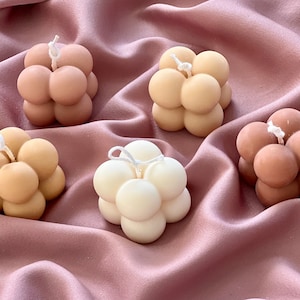 Peut inclure: Quatre bougies en forme de bulles de différentes couleurs : marron, beige et blanc. Les bougies sont disposées sur un tissu satiné rose.