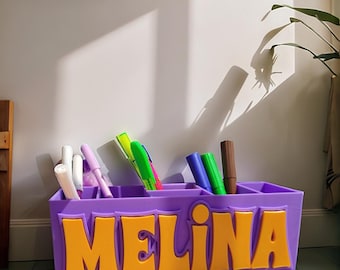 May include: A rectangular purple plastic desk organiser featuring the name MELINA in large, raised yellow letters on the front. The organiser has multiple compartments filled with various coloured markers, including white, purple, green, and brown.