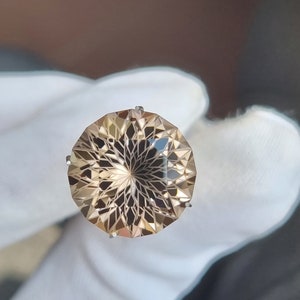 Può includere: Una pietra preziosa rotonda, sfaccettata, di colore marrone chiaro con un motivo a stella al centro. La pietra preziosa è incastonata in una montatura d'argento.