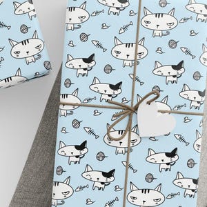 May include: Gift-wrapped presents with light blue paper featuring a repeating pattern of cartoon cats, fish bones, and yarn balls. The gifts are tied with twine and have heart-shaped tags. A ball of twine sits nearby.