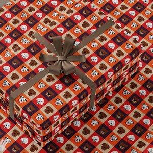 Könnte beinhalten: Orangefarbenes, braunes und weißes Geschenkpapier mit einem sich wiederholenden Muster aus Hundegesichtern. Das Papier ist um eine Geschenkbox mit einem braunen Band gewickelt.