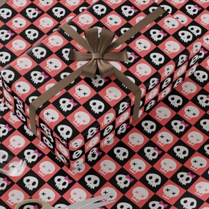 May include: A wrapped gift box with a brown ribbon sits on wrapping paper. The paper features a repeating pattern of white skulls with pink bows and crosses on a red and black checkered background. Scissors are in the foreground.