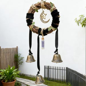 May include: A handcrafted wreath with a natural twig base, accented with black velvet, green leaves, and a gold crescent moon and star charm. Black ribbons and three brass bells hang below, creating a whimsical, decorative piece.