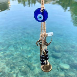 Puede incluir: Un amuleto de ojo turco de cristal azul con iris blanco, suspendido de un cordón de fibra natural. Debajo, una colección de pequeños amuletos incluye una llave, una luna creciente y cuentas, todos en tonos bronce. Una pequeña campana cuelga en la parte inferior.