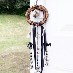 May include: A handcrafted, circular grapevine wreath with black ribbon and hanging charms. Charms include keys, bells, a bat, and a sun design. The wreath is adorned with a small glass bottle and a miniature hat. The wreath hangs from a white door.