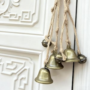 Puede incluir: Un carillón decorativo con seis campanas de latón colgando de una cuerda de yute trenzada. Las campanas están suspendidas en un grupo, con una mezcla de formas. El fondo es una puerta blanca con detalles tallados.
