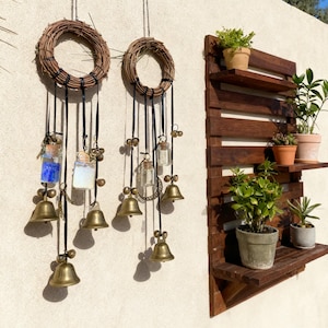May include: Two hanging grapevine wreaths with black ribbons, glass bottles, and brass bells. A wooden shelf with potted plants is mounted on the wall. The scene is set against a white wall and a bright blue sky.