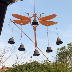 May include: A metal dragonfly wind chime with six bronze bells suspended from chains. The dragonfly is a rusty orange colour and hangs from a chain. The background features green foliage and a blue sky.