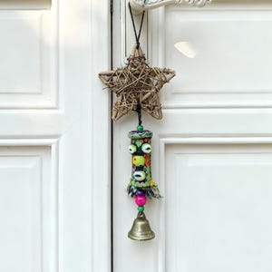 Könnte beinhalten: Ein dekorativer Türhänger mit einem geflochtenen Stern, einem bunten zylindrischen Ornament mit Perlenakzenten und einer kleinen Messingglocke. Die Tür ist weiß mit einem dekorativen Griff.