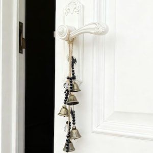 Puede incluir: Un adorno de puerta decorativo con pequeñas campanas de latón, cuentas negras y cristales transparentes cuelga de un tirador de puerta blanco. La puerta y el tirador son blancos, con un fondo oscuro.