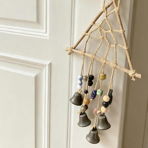 Puede incluir: Un carillón hecho a mano con un marco triangular de madera y un diseño de telaraña tejida. El carillón presenta pequeñas campanas grises y cuentas de colores colgando de hilo de yute. Está suspendido de un pomo de puerta blanco.