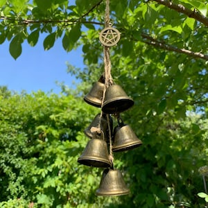 Puede incluir: Un grupo de pequeñas campanas de latón antiguo colgadas de un cordón de hilo natural. Un amuleto de pentáculo dorado está en la parte superior. Las campanas cuelgan de una rama de árbol sobre un fondo de follaje verde y cielo azul.
