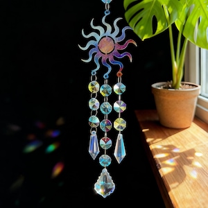 Puede incluir: Un atrapasoles decorativo con un colgante en forma de sol con un acabado iridiscente. Debajo del sol, una cadena sostiene varias cuentas y colgantes de cristal transparente y facetado. El atrapasoles cuelga sobre un fondo oscuro.