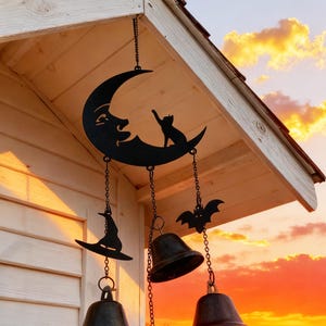 Puede incluir: Carrillón de Halloween de metal negro con una luna creciente con una cara y una silueta de gato. El carillón incluye un sombrero de bruja, un murciélago y tres campanas. El fondo muestra una puesta de sol sobre un lago.