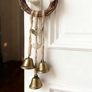 Peut inclure: Une couronne rustique en brindilles brunes tressées, ornée de trois cloches en laiton suspendues à de la ficelle de jute. Un charm papillon décoratif et une poignée de porte blanche sont également visibles. La couronne est accrochée à une porte blanche.