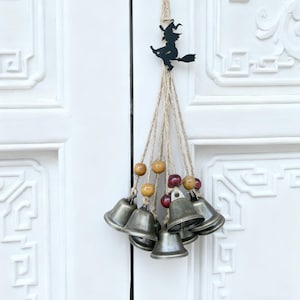 May include: A Halloween-themed door hanging featuring a black silhouette of a witch on a broom. The witch is suspended from a natural twine loop, with multiple strands of twine holding small silver bells, wooden beads, and red beads.