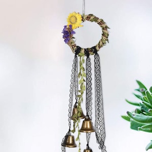 Może przedstawiać: Dekoracyjny wieniec z suszonymi kwiatami i zielenią, ozdobiony czarnymi wstążkami z koronki i czterema złotymi dzwonkami.