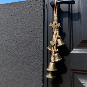 Könnte beinhalten: Eine Halloween-Türdekoration mit kleinen Messingglocken und einem Metallanhänger in Form einer Fledermaus und eines Geistes. Die Glocken hängen an einem Naturseil und sind an einer schwarzen Tür mit einem schwarzen Griff befestigt.