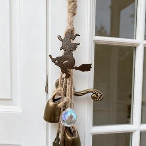 Könnte beinhalten: Halloween-Türdekoration. Eine bronzefarbene Hexe auf einem Besen hängt an einem Naturseil. Unter der Hexe befinden sich drei bronzefarbene Glocken, ein Kristallprisma und ein bronzefarbenes Kreuz. Die Tür ist weiß mit einem dekorativen Griff.