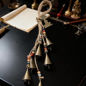 Peut inclure: Un carillon décoratif avec de petites cloches en laiton antique suspendues à une corde de jute. Le carillon comprend des perles en bois colorées et une silhouette métallique d'un chien. Les cloches sont en forme de cône et pendent à différentes longueurs.