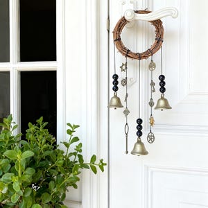 May include: A decorative wind chime with a woven wreath base, hanging from a white door handle. The chime features black beads, gold-colored bells, and small metal charms, including keys and stars. The door is white, and a green plant is visible.