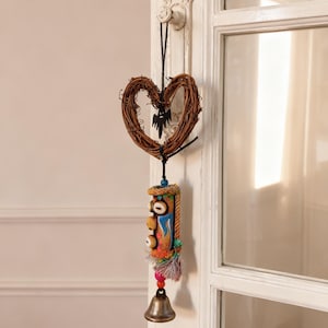 Puede incluir: Adorno colgante decorativo. Un diseño en forma de corazón de vid trenzada con una silueta negra cuelga de una puerta blanca. Debajo, una pieza de madera tallada colorida con una campana y borla cuelga de un cordón negro.