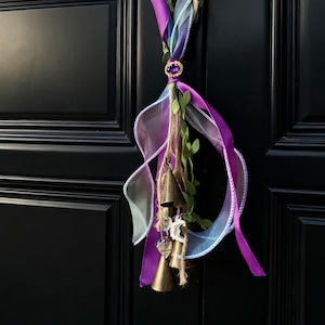 May include: A decorative door hanger with purple and sheer ribbons, faux greenery, and small brass bells. A jeweled clasp secures the ribbons. The door is black.
