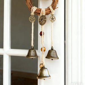 May include: A rustic wreath made of woven twigs, adorned with hanging bronze bells and star-shaped charms. The wreath is suspended from a white door handle, creating a charming, natural accent for home decor.