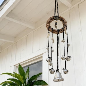 Könnte beinhalten: Ein hängendes Windspiel mit einem kreisförmigen geflochtenen Kranz oben. Schwarze Schnüre gehen herab, verziert mit silbernen Glocken und dekorativen Metallanhängern. Das Windspiel hängt an einer schwarzen Halterung an einer weißen Holzdecke.