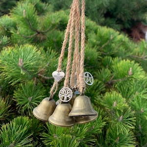 Könnte beinhalten: Ein hängender Windspiel mit drei Bronzeglocken und dekorativen Anhängern. Die Anhänger umfassen ein Herz mit dem Schriftzug "Blessed Be", einen Lebensbaum und ein Pentagramm. Das Windspiel ist an einem Naturseil und einer Bronzeglocke aufgehängt.