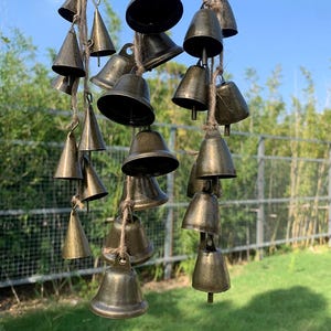 May include: A collection of bronze-colored metal bells of varying sizes, suspended from natural twine. The bells are arranged in a cascading pattern, creating a wind chime effect. The background features green grass and foliage.