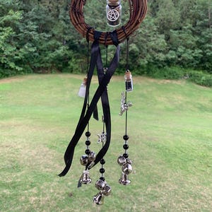 Könnte beinhalten: Ein handgefertigter Traumfänger mit einer gewebten braunen Kranzbasis. Er verfügt über kleine Glasfläschchen, schwarze Bänder, silberne Glocken und dekorative Anhänger. Der Traumfänger hängt im Freien vor dem Hintergrund grüner Bäume und Gras.