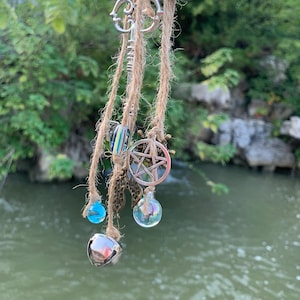 May include: A hanging decorative piece featuring silver bells, charms, and beads suspended from jute twine. Charms include a key, a pentagram, and a heart with wings. The background shows a body of water and green foliage.