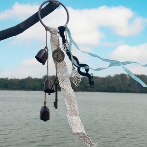 Könnte beinhalten: Ein Traumfänger aus weißer Spitze mit einem Holzring, schwarzen und blauen Bändern und drei kleinen Glocken, die daran hängen. Der Traumfänger hängt an einem Ast vor einem Hintergrund aus blauem Himmel und Wasser.