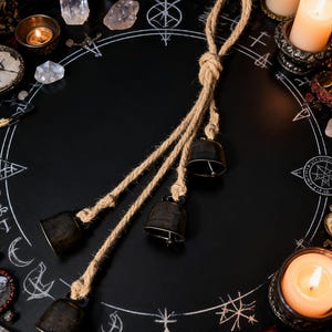 Könnte beinhalten: Drei dunkle Metallglocken, die an einem Naturseil hängen, vor einem schwarzen Hintergrund mit weißen Kreidesymbolen. Die Glocken haben ein rustikales, gealtertes Aussehen. Die Szene ist von Kerzen, Kristallen und einem Schädel umgeben, was ein mystisches oder spirituelles Thema suggeriert.